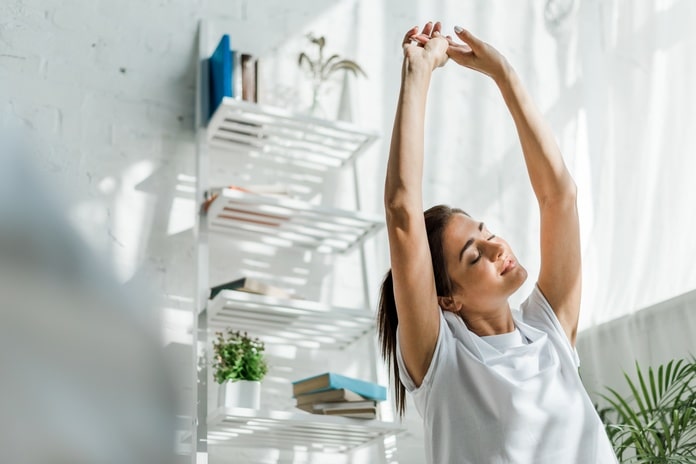 A young woman stretching her arms over her head and gently leaning to one side as natural light streams in through a window.
