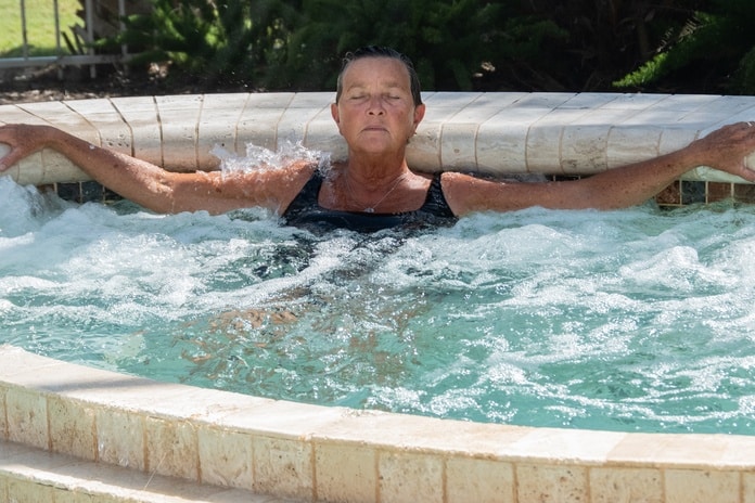 A senior woman enjoying her in-ground hot tub at home. Her eyes are relaxed and shut and her arms are stretched out.