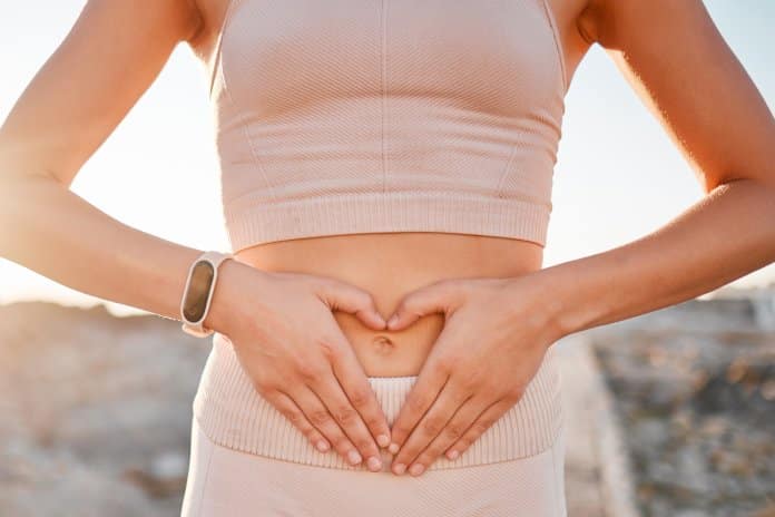 A woman wearing an athleisure set places her hands on her stomach to make a heart with the sun shining behind her.