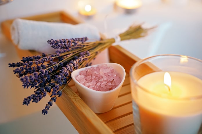 A wooden bathtub tray over a bath holds a candle, lavender, a hand towel, and a small bowl of pink Epson salt.