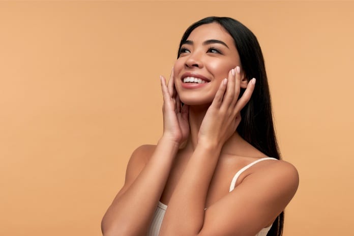 A woman wearing a white tank top standing in front of a beige background while holding her hands to her face and smiling.