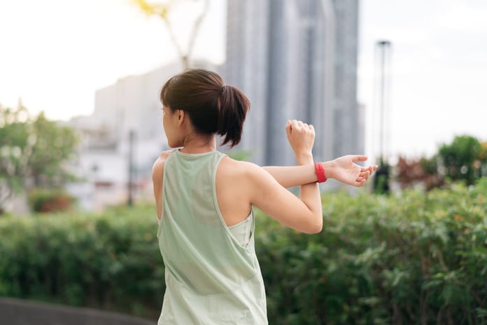 A woman wearing a light green tank top and red watch stretches her arms outside while surrounded by green shrubs.