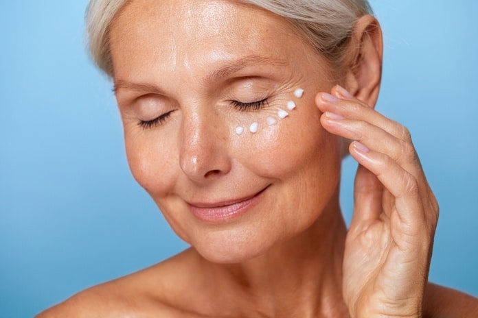 An older woman with a creamy facial moisturizer dotted underneath her eye. She is smiling with her eyes closed.