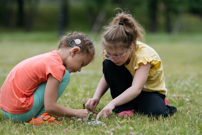Two children kneeling in a grassy field to inspect things on the ground. They both have their hair up in a style.