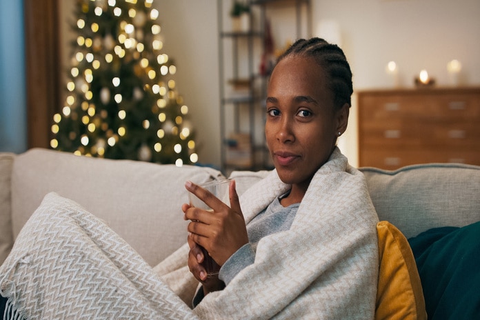 A woman sits on a couch, is wrapped in a blanket, and holds a mug. A Christmas tree is in the background.
