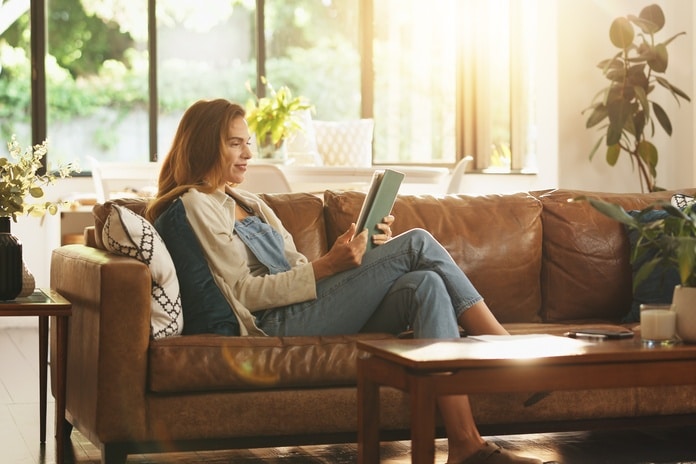 A woman sits on a brown leather couch and looks at a tablet. Sun shines through the window, and potted plants adorn the room.