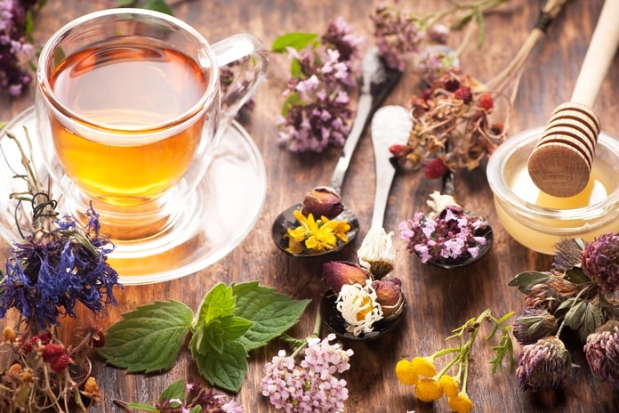A clear mug of hot tea next to a container of honey with a honey dipper resting on it. Various dried herbs are scattered about.