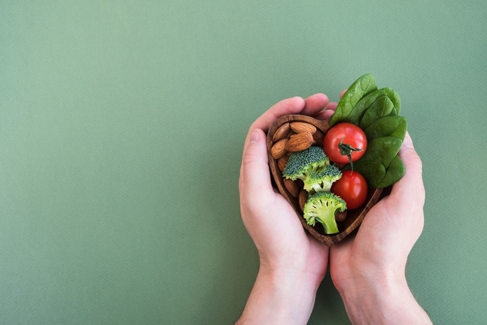 Hands holding a plate of heart-healthy foods with greens, vegetables, and nuts on a bright green background.
