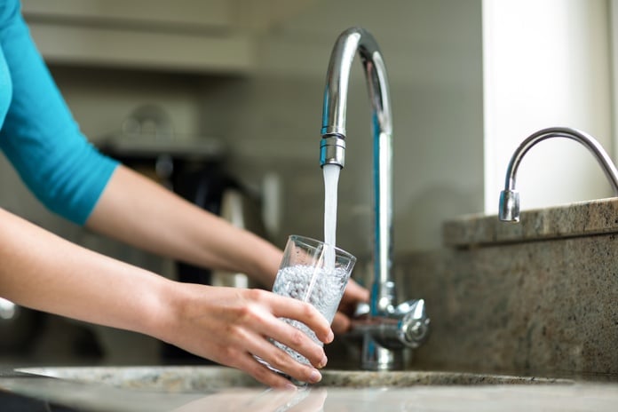 A close-up of a woman holding a tall glass up to a kitchen sink faucet and filling it with tap water.
