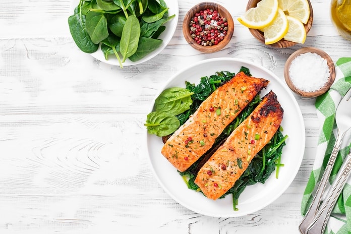Grilled salmon fillets on sautéed spinach with lemon slices, peppercorns, and sea salt on a light wooden surface.