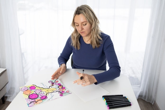 A woman sitting in front of a table with a half finished picture on it. The woman is coloring with pens.