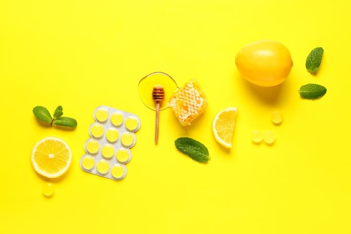 Several throat lozenges with honey, mint leaves, and lemon slices arranged on a bright yellow background.