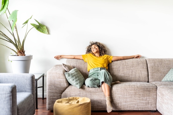 A woman in a yellow shirt sitting on a sofa in a neutral-colored living room. She is smiling with her head back and her arms out.