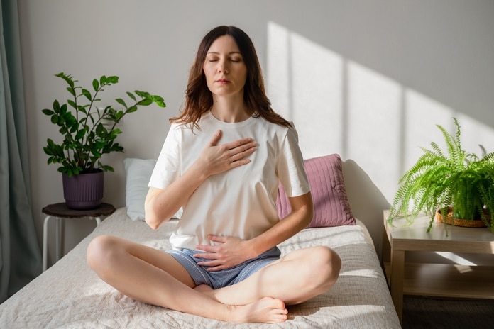 A close-up of a woman sitting on a bed with her eyes closed, legs crossed, one hand on her chest, and one hand on her belly.
