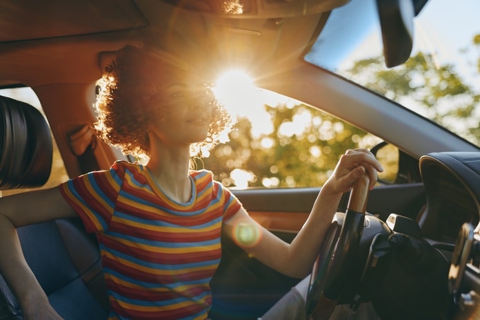 A woman wearing a striped shirt sits in the driver's seat of a car as the sun sets behind the trees.